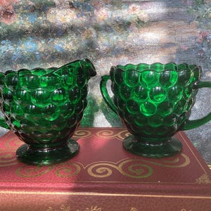 May include: A pair of emerald green glass vessels featuring a raised hobnail texture. One piece is a creamer with a pouring spout and a single handle, while the other is a sugar bowl with two side handles. Both items sit on circular pedestal bases.