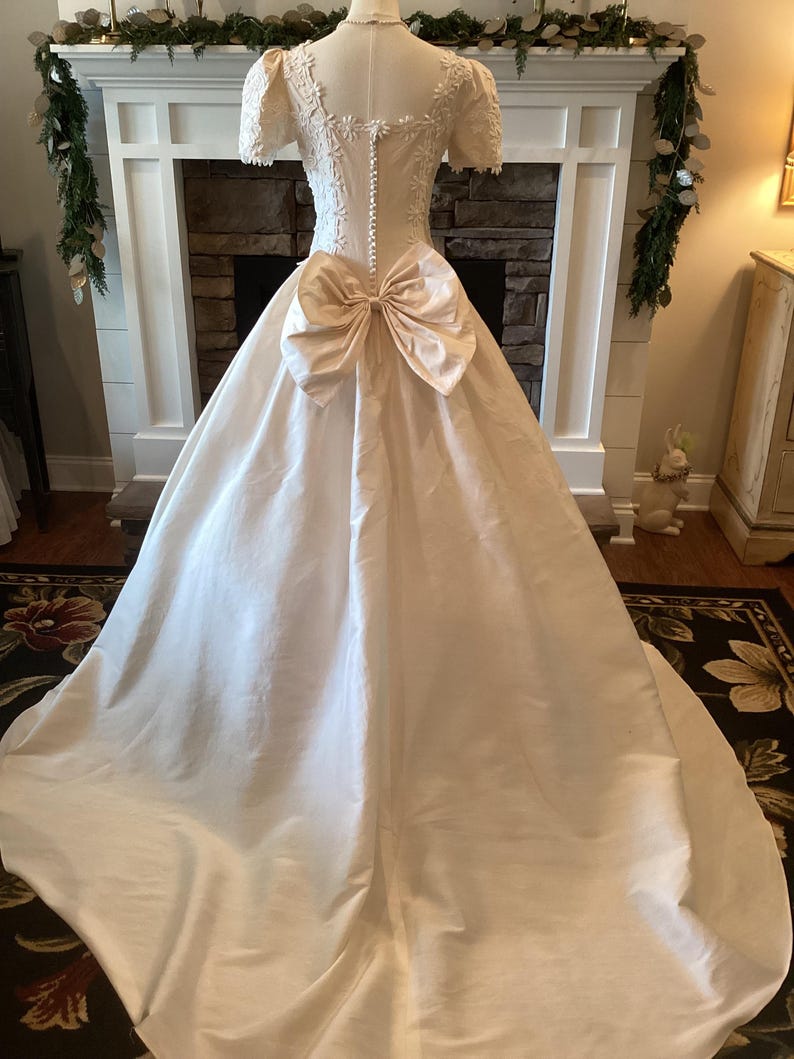 Op de afbeelding: Een vintage ivoorkleurige trouwjurk met een grote strik op de rug. De jurk heeft korte mouwen met bloemenkantdetails, een getailleerd lijfje en een lange, vloeiende rok. De achterkant heeft een rij kleine knopen.