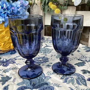 May include: Two cobalt blue stemmed water glasses with a faceted design. The glasses sit on a floral patterned tablecloth. A yellow vase with blue flowers is in the background.