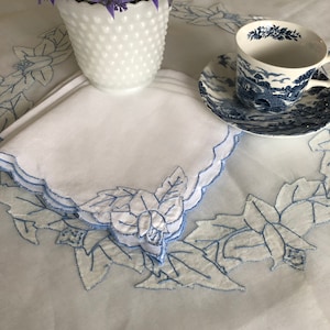 May include: White embroidered linen napkins with blue floral designs. The napkins are folded and stacked on a white tablecloth with blue embroidered floral designs. A white milk glass vase with lavender flowers and a blue and white teacup and saucer are in the background.
