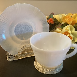 May include: A white milk glass teacup and saucer set with a delicate floral design. The set is displayed on a gold metal stand.
