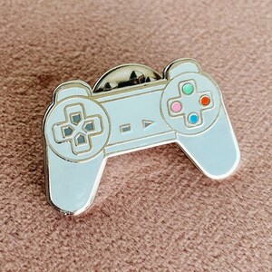 Peut inclure: Une épingle en émail argentée en forme de manette de jeu vidéo. La manette a un fond blanc avec des boutons noirs et une plaque frontale grise. Les boutons sont de couleur rouge, bleu, vert et rose.