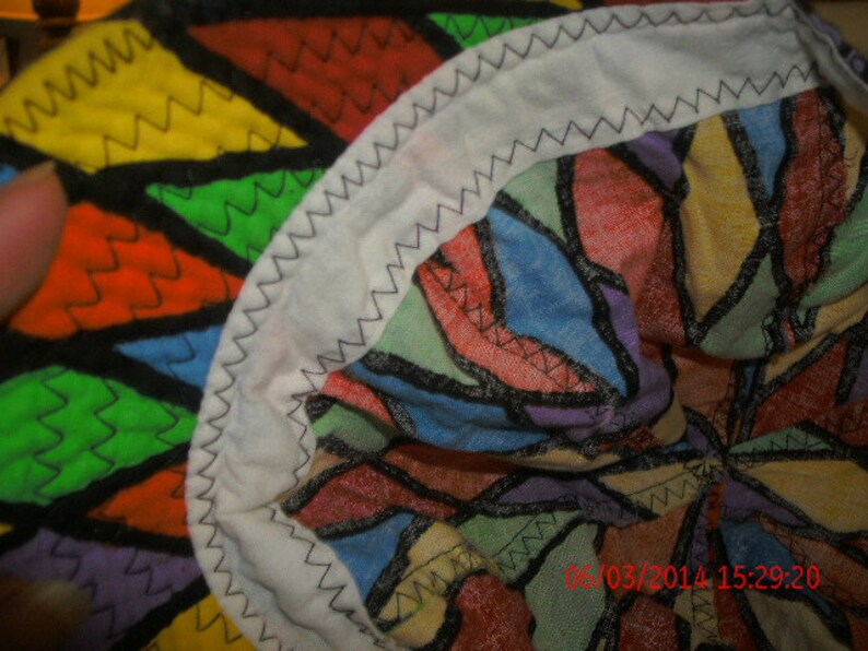 K&ouml;nnte beinhalten: Ein farbenfroher Stoff mit einem geometrischen Muster in Rot-, Gelb-, Gr&uuml;n-, Blau- und Violettt&ouml;nen. Der Stoff hat einen wei&szlig;en Rand mit einer Zickzacknaht.