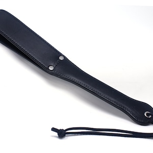 May include: A black leather paddle with a stitched edge and a metal ring at the end. A black cord is attached to the ring.