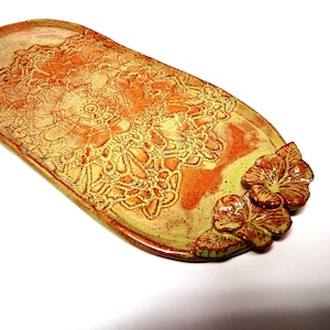 May include: An elongated oval ceramic tray featuring a vibrant orange and yellow glaze with a speckled finish. The surface is embossed with a detailed lace-like pattern and two sculpted three-dimensional flowers attached to one end.