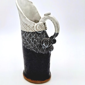 May include: A tall ceramic pitcher featuring a two-tone design with a white upper section displaying a grid pattern and a black lower section with textured detailing. The handle is adorned with a spiral accent, and the base reveals a natural clay color.