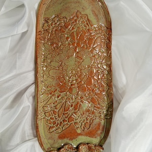 May include: An elongated oval ceramic tray featuring a speckled sage green and burnt orange glaze. The surface displays a raised, intricate lace-like texture pattern. Three sculpted ceramic flowers are attached to the bottom edge of the tray.