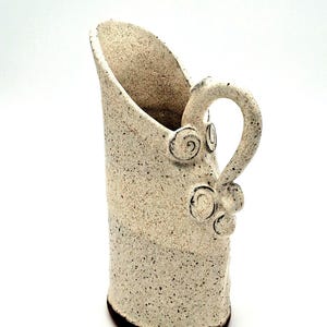 May include: A tall, off-white ceramic pitcher with a speckled texture throughout. The vessel features a slanted rim, a curved handle attached to the side, and decorative spiral clay accents near the handle base. The bottom edge is dark brown.