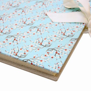 Könnte beinhalten: Ein gebundenes Notizbuch mit hellblauem Einband, verziert mit einem floralen Muster aus weißen Blüten und roten Akzenten. Das Notizbuch ist mit einem cremefarbenen Band gebunden. Die Kanten des Journals sind in einem neutralen Beige gehalten.