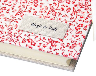May include: A hardcover book with a white cover featuring a red floral pattern. A rectangular label with the text "Birgit & Ralf" is attached to the cover. The book's spine is visible, and a white ribbon is attached.