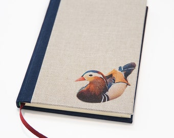 May include: A hardback notebook featuring a beige linen-textured cover with a dark blue spine and a vibrant illustration of a mandarin duck. The notebook includes a thin red ribbon bookmark extending from the bottom edge of the binding.