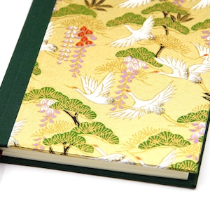 Op de afbeelding: Een hardcover notitieboek met een gouden omslag met witte kraanvogels, groene dennenbomen en paarse blauweregen. De rug is donkergroen en een bordeauxrood lint als bladwijzer is zichtbaar. Het notitieboek is gesloten.