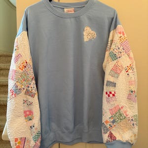 May include: A light blue crewneck sweatshirt featuring long sleeves made from a multicolored patchwork quilt fabric. A heart-shaped patch crafted from the same patchwork quilt material is stitched onto the upper left chest area.