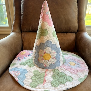 May include: A conical hat constructed from quilted fabric featuring a hexagonal patchwork pattern. The design incorporates floral and gingham prints in soft shades of pink, sage green, light blue, and yellow, with visible stitching.