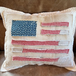May include: A square decorative pillow with a rustic design. The pillow features a blue rectangle with white stars in the upper left corner, and red and white striped accents, resembling the American flag. The pillow has a cream-colored background and a textured appearance.