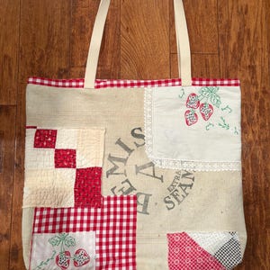 May include: A patchwork tote bag with cream-colored fabric and red gingham and checkered patterns. The bag features embroidered strawberries and a cream-colored handle. The top edge is trimmed with red and white gingham.