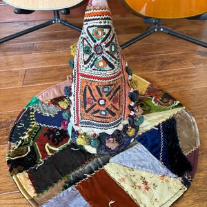 May include: A conical textile sculpture featuring intricate embroidery, small circular mirrors, and colorful pom-poms, resting on a circular base made of various patchwork fabric scraps including velvet, floral patterns, and dark textured materials.