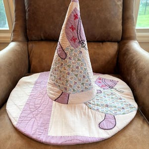 May include: A conical hat constructed from quilted fabric featuring a patchwork design. The hat displays a light blue floral pattern, solid lavender sections, and white panels. It is accented with embroidered red flower motifs and black outlines.