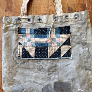 May include: A gray canvas tote bag with a patchwork design. The patchwork features blue, pink, and black fabric squares. The bag has reinforced grommets and sturdy handles. The bag has a drawstring closure.