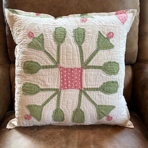 May include: A square throw pillow featuring a white quilted fabric base with a central pink square patch. The design includes green fabric flower-like shapes with small pink accents arranged in a radial pattern, all finished with detailed white stitching.