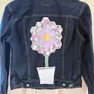 May include: Dark blue denim jacket with a large, colorful flower applique on the back. The flower features a lavender center, yellow center, and pink and white petals. The jacket has button closures and pockets.