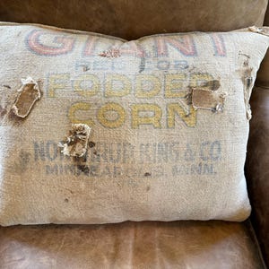 May include: A rectangular throw pillow made from a tan burlap sack featuring the text GIANT RED TOP FODDER CORN and NORTHRUP KING & CO. MINNEAPOLIS, MINN. The fabric has a coarse, woven texture with visible frayed patches and dark spots.