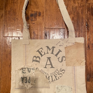 May include: Cream-colored tote bag with a distressed aesthetic. The bag features a large circular graphic that reads "Bemis A Extra Heavy Seamless." Patches of fabric are sewn onto the bag. The bag has a long strap.