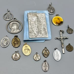May include: A collection of silver and gold religious pendants, including a cross, a heart, and a variety of images of the Virgin Mary. The pendants are arranged on a light grey surface.
