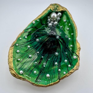 May include: A decorative oyster shell painted in vibrant green with a metallic gold rim. The interior features black ink-like streaks radiating from the center, accented with numerous small white pearls and one larger central pearl at the top.