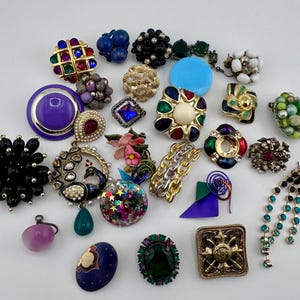 May include: A selection of vintage earrings and brooches in diverse designs and hues. Includes clip-on earrings, drop earrings, and pins with rhinestones, pearls, and enamel. Features gold, silver, and jewel-toned accents.