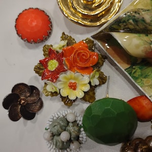 May include: A selection of vintage brooches and decorative items. Includes a bright orange faceted brooch, a bronze flower brooch, and a floral brooch with orange and yellow flowers. Also, a green geometric sphere.