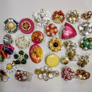 May include: A selection of vintage brooches and earrings in diverse designs and colours. Features floral, geometric, and abstract styles with pearl, rhinestone, and enamel embellishments. The jewellery is displayed on a white surface.