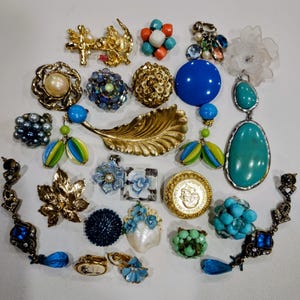 May include: A selection of vintage jewellery items, including earrings, brooches, and pendants. The jewellery features various designs, colours, and materials, such as gold, blue, turquoise, and pearls. The pieces are arranged on a white surface.