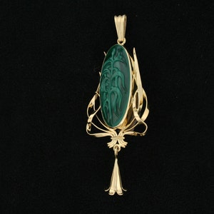 Puede incluir: Un colgante de oro con una piedra de malaquita verde tallada con un dise&ntilde;o floral. El colgante est&aacute; suspendido de una cadena delicada.