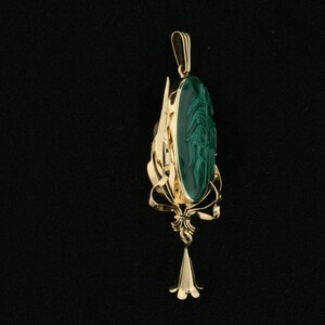 Puede incluir: Un colgante de oro con una piedra de malaquita verde tallada con un dise&ntilde;o. El colgante est&aacute; unido a una cadena de oro.