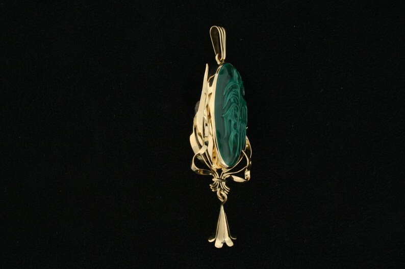 Puede incluir: Un colgante de oro con una piedra de malaquita verde tallada con un dise&ntilde;o. El colgante est&aacute; unido a una cadena de oro.
