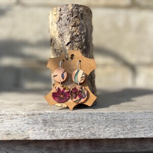May include: Round floral earrings with silver hooks. The earrings feature a light tan background with a burgundy and green floral pattern. The earrings are displayed on a brown cardboard tag and a wooden log.