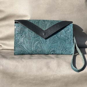 Pode incluir: Uma carteira de mão de couro azul turquesa com uma aba de couro preto e um design em relevo floral. A carteira de mão tem uma alça de pulso.