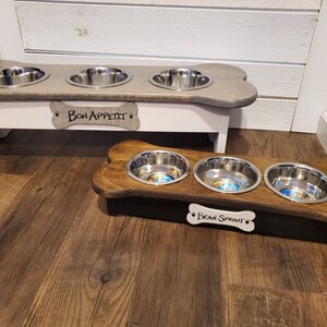 May include: Two wooden dog bowl stands with three stainless steel bowls each. The top stand is white with a gray bone-shaped top and the text "Bon Appetit" on a bone-shaped tag. The bottom stand is brown with a black base and the text "Bean Sprout" on a bone-shaped tag.