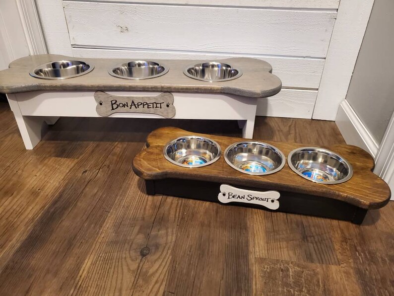 May include: Two wooden dog bowl stands with three stainless steel bowls each. The top stand is white with a gray bone-shaped top and the text "Bon Appetit" on a bone-shaped tag. The bottom stand is brown with a black base and the text "Bean Sprout" on a bone-shaped tag.