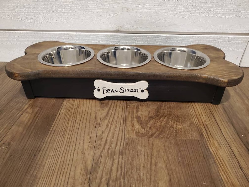 May include: A wooden dog bowl stand with three stainless steel bowls. The stand is shaped like a bone and has a black base with a white bone-shaped plaque that says "Bean Sprout".