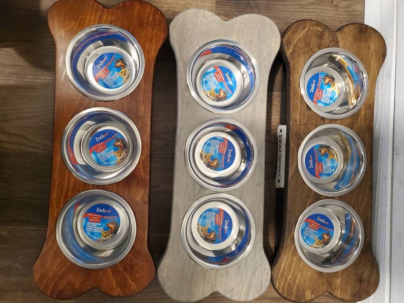 May include: Three bone-shaped wooden dog food bowls with stainless steel bowls. The bowls are in different colors: brown, gray, and brown. The bowls are labeled "Indigrow".