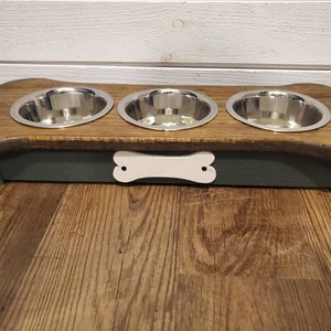 May include: A wooden dog bowl stand with three stainless steel bowls. The stand is shaped like a bone and has a white bone cutout on the front. The stand is painted dark green and has a natural wood finish on top.