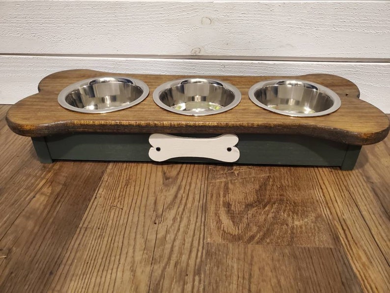 May include: A wooden dog bowl stand with three stainless steel bowls. The stand is shaped like a bone and has a white bone cutout on the front. The stand is painted dark green and has a natural wood finish on top.
