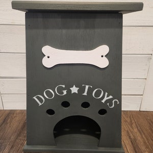 Może przedstawiać: Drewniana skrzynka na zabawki dla psa w ciemnoszarym kolorze z wyciętym białym kością i napisem "DOG TOYS" białymi literami. Skrzynka ma wycięcie w kształcie odcisku łapy z czterema otworami.