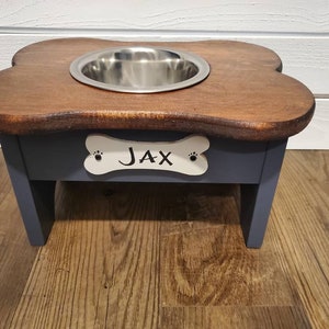 May include: A wooden dog bowl stand with a bone-shaped cutout and the name "JAX" painted on it. The stand is painted a dark blue color and has a stainless steel bowl.