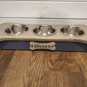May include: A wooden dog bowl stand in the shape of a bone with three stainless steel bowls. The stand is painted a light gray color and has the name "Ranger" engraved on the front.