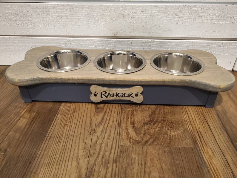 May include: A wooden dog bowl stand in the shape of a bone with three stainless steel bowls. The stand is painted a light gray color and has the name "Ranger" engraved on the front.