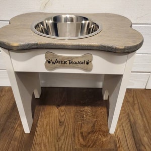May include: White wooden dog food and water stand with a bone-shaped top and a stainless steel bowl. The stand has a sign that says "Water Trough" with paw prints.