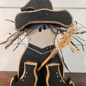 May include: Wooden Halloween witch figurine with a black hat, boots, and outfit. The witch has a straw broom and a light-colored face with two black eyes. The hat and boots have white speckles.